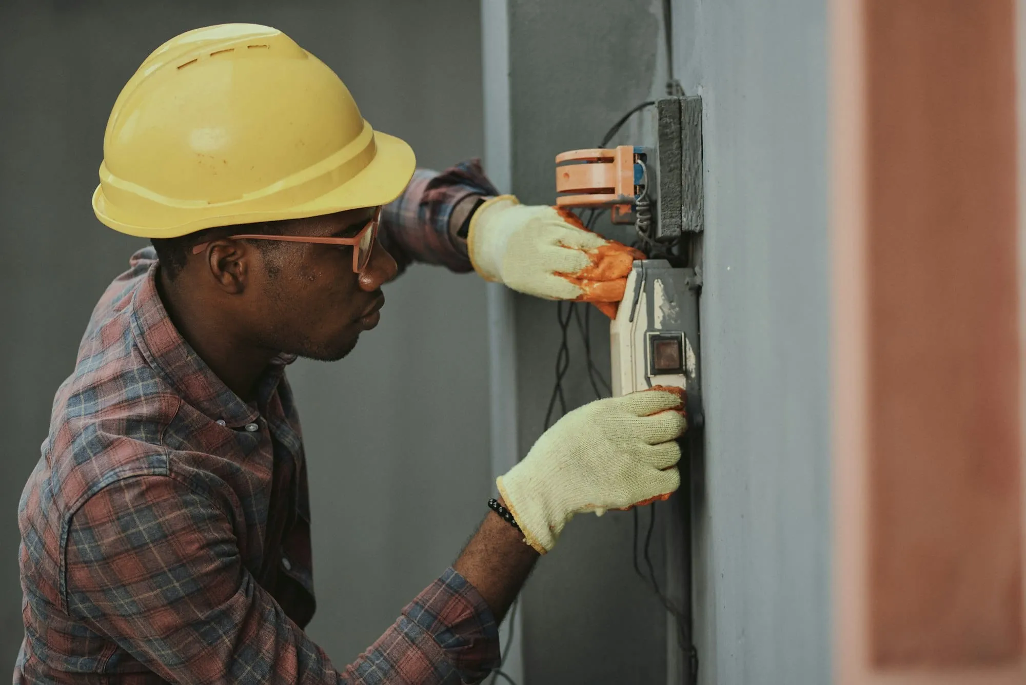A maintenance engineer in a hard hat and gloves working on a wall-mounted electrical unit.