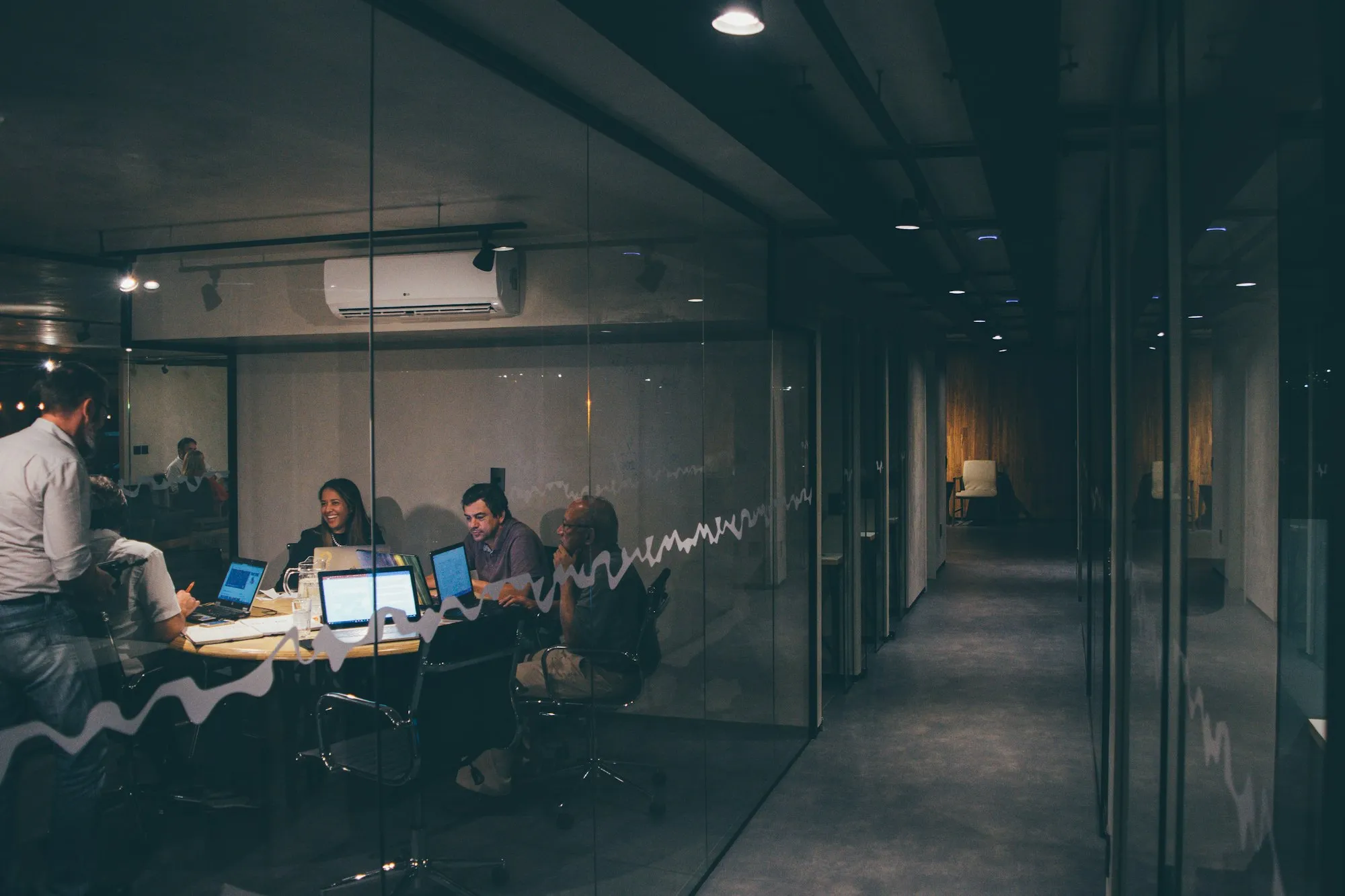 A team around a table in a glass-walled meeting room, laptops open