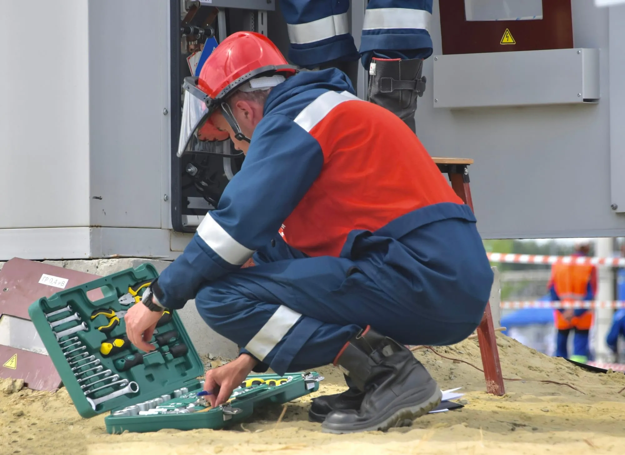 An engineer in overalls and a hard hat crouching over an open tool case on site.